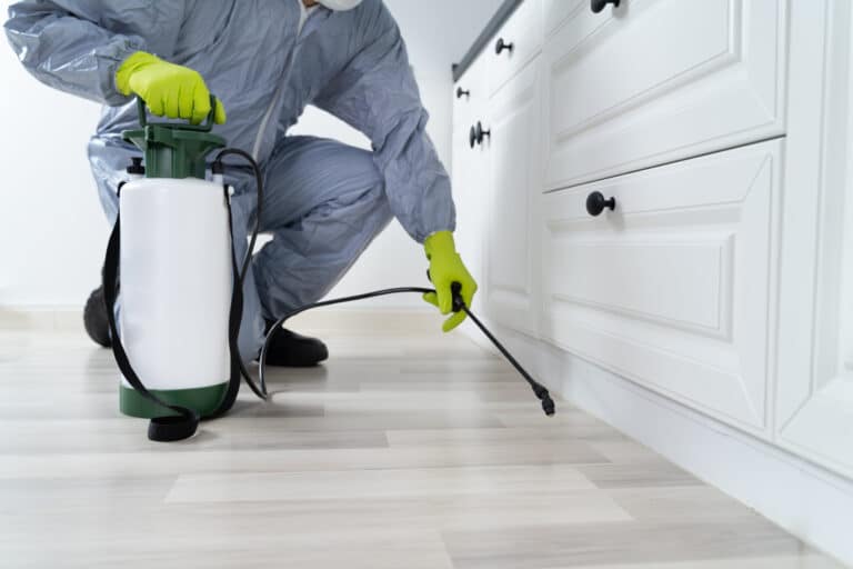 Exterminator man in proper gear spraying pesticide onto a light grey and tan floor with white cabinet drawers